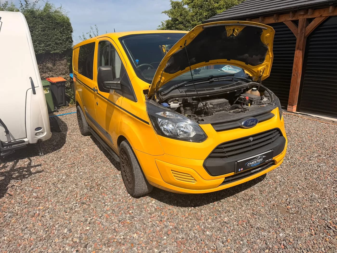 A yellow Ford Transit Custom with its bonnet up on a gravel driveway during a mobile Chester Tuning visit